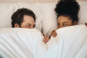 A couple waking up together remaining tucked in the covers of the bed.