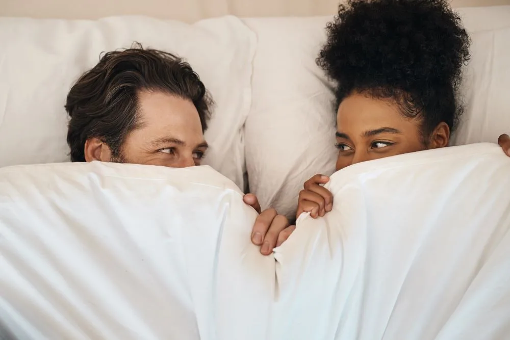 A couple waking up together remaining tucked in the covers of the bed.