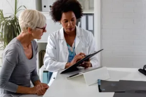 A female doctor sits at her desk and chats to an elderly female patient while looking at pre-appointment prepa.