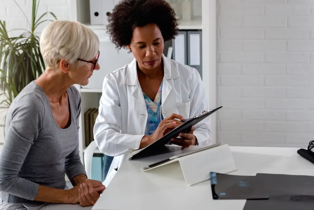 A female doctor sits at her desk and chats to an elderly female patient while looking at pre-appointment prepa.
